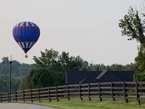 A colorful hot air balloon floats above a house, with a wooden fence and trees in the background.
