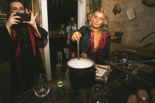 A woman stirs a cauldron with smoke, while another person takes a photo in a dimly lit, atmospheric setting.