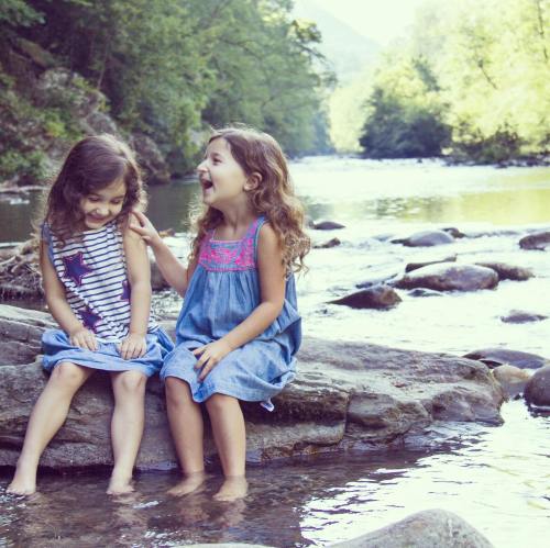 Two young girls sit on rocks by a river, laughing and enjoying a sunny day surrounded by trees.