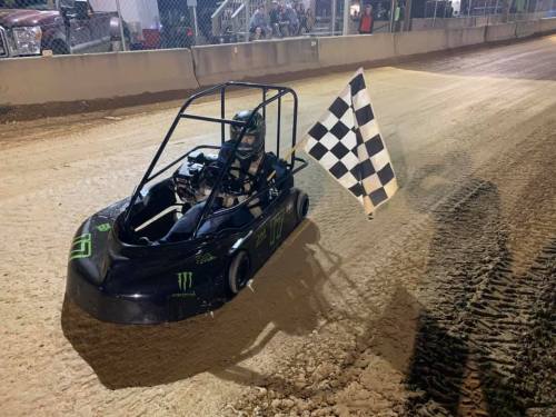 A go-kart driver holds a checkered flag while racing on a dirt track at night.