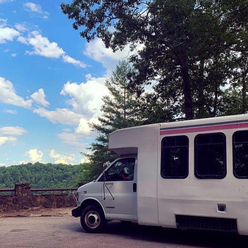 A white bus parked near a scenic view of trees and a blue sky with fluffy clouds.