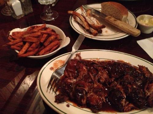 A plate of barbecue meat with sauce, served with sweet potato fries and a side of bread on a wooden table.