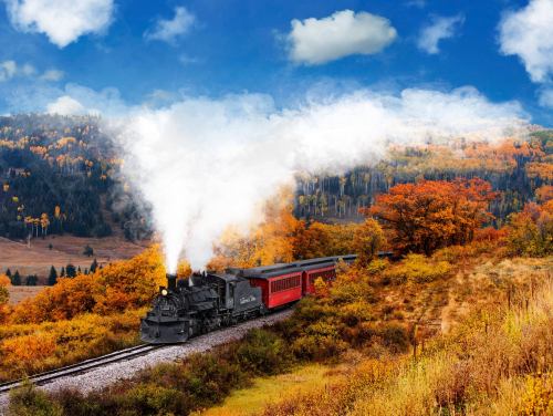 A vintage steam train travels through a colorful autumn landscape with vibrant trees and a blue sky.