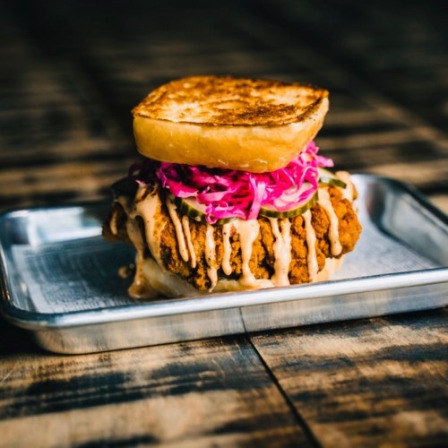 A crispy fried chicken sandwich with pickled red cabbage and drizzled sauce, served on a toasted bun.
