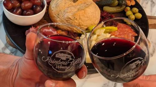 Two hands clink wine glasses filled with red wine, with a charcuterie board and snacks in the background.