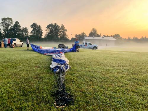 A hot air balloon crew prepares the balloon on a grassy field at sunrise, with mist in the background.