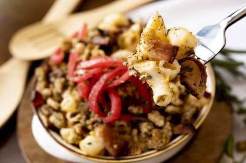 A fork holding a bite of macaroni and cheese topped with crispy bacon and pickled onions, served in a bowl.