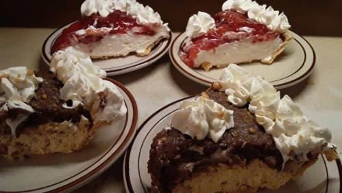 Four slices of pie on plates, featuring whipped cream and fruit toppings, with one slice showing a chocolate layer.