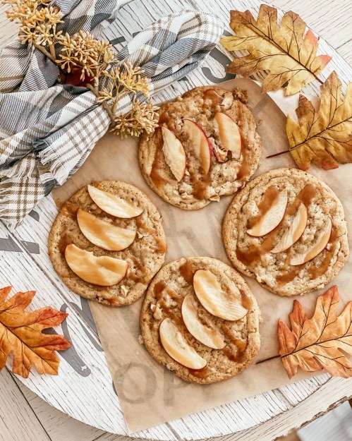 Four cookies topped with apple slices and caramel, surrounded by autumn leaves and a plaid napkin on a wooden tray.