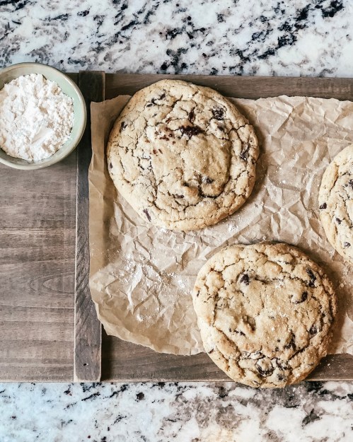 Three large chocolate chip cookies on parchment paper, with a bowl of flour nearby, on a wooden surface.