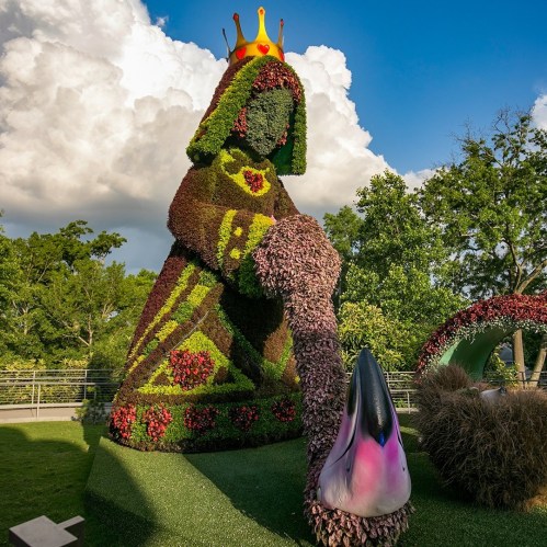 A large floral sculpture of a queen holding a flower, surrounded by greenery and a blue sky with clouds.