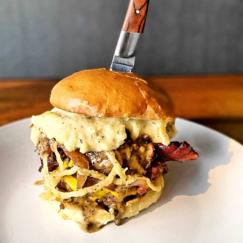 A juicy burger topped with creamy sauce, crispy onions, and bacon, served on a toasted bun with a knife inserted.
