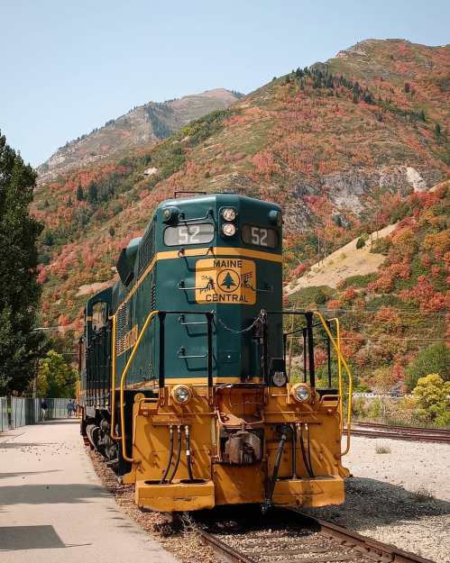A vintage train in green and yellow parked beside a scenic mountain landscape with vibrant autumn foliage.