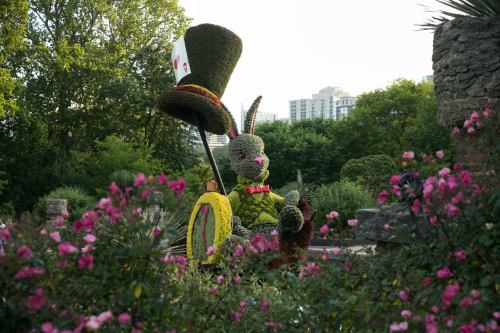 A topiary rabbit in a hat sits among pink flowers, with greenery and city buildings in the background.