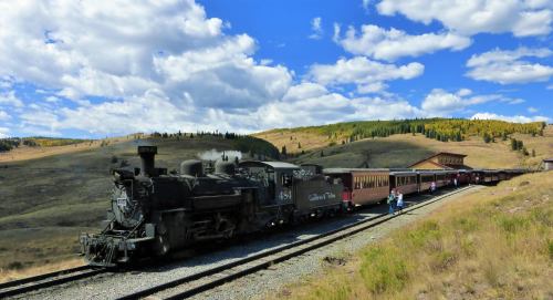 A vintage steam train stops at a scenic station surrounded by hills and trees under a blue sky with fluffy clouds.