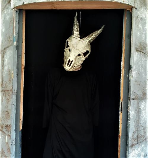 A figure in a black outfit stands in a doorway, wearing a horned skull mask. The setting has a dark, eerie atmosphere.