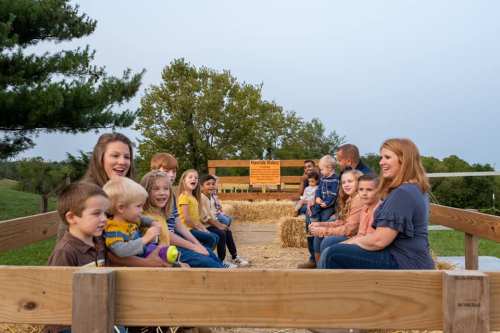 A group of children and adults sit on a hayride, smiling and enjoying a sunny day outdoors.
