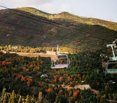 A ski lift chair hangs over a colorful autumn landscape with mountains and trees in vibrant fall foliage.