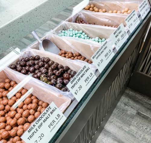 A display of various chocolate malt balls in bins, labeled with prices per pound.