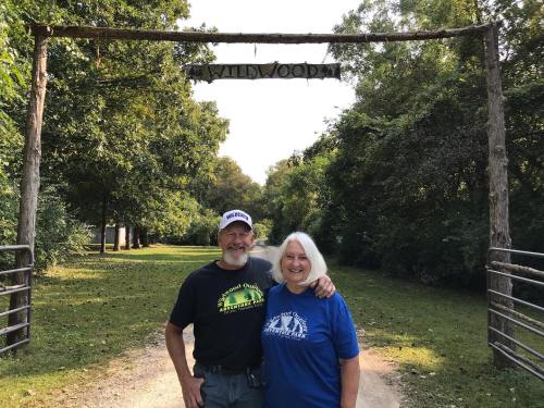 A smiling couple stands under a wooden sign that reads "Wildwood," surrounded by trees and a gravel path.