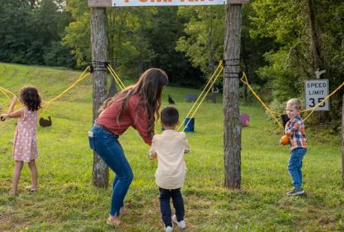 Children and a woman play at a pumpkin patch, pulling on ropes with pumpkins in a scenic outdoor setting.