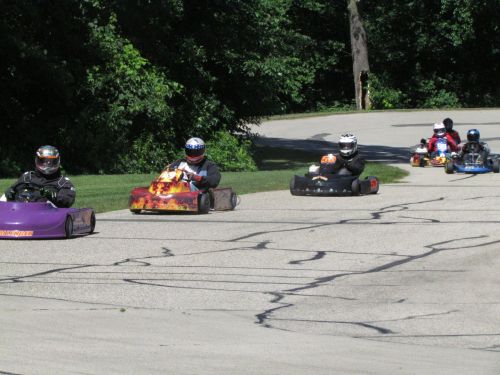 A group of go-kart racers in helmets compete on a winding track surrounded by greenery.