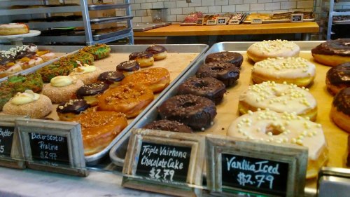 A display of assorted donuts, including chocolate, vanilla, and specialty flavors, with price tags on a wooden tray.