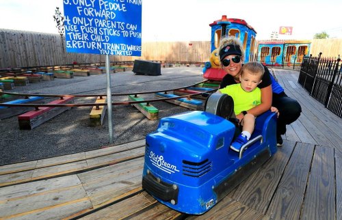 A woman and a child enjoy a ride on a blue toy train at an amusement park, with colorful tracks in the background.
