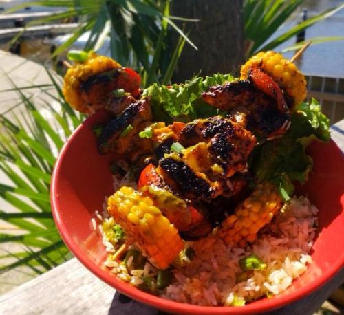 A vibrant bowl of grilled shrimp skewers with corn, served on a bed of rice and lettuce, set against a sunny backdrop.