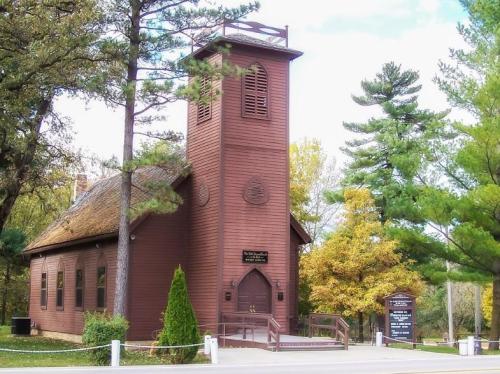A rustic brown church with a tall bell tower, surrounded by trees and autumn foliage.