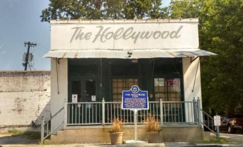 A vintage building with a sign reading "The Hollywood" and a historical marker outside, surrounded by greenery.