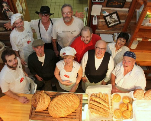 A group of bakers poses together in a bakery, surrounded by various freshly baked breads on display.