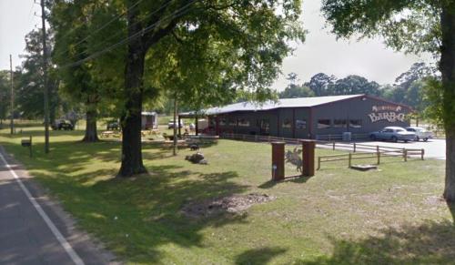 A barbecue restaurant with a gray building, surrounded by trees and a grassy area, located by a road.