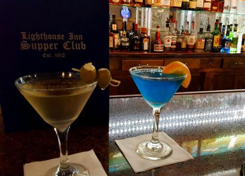 Two colorful cocktails on a bar counter, with a menu in the background and various bottles of liquor displayed.