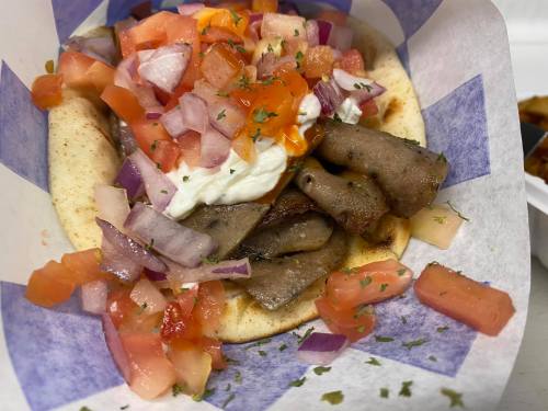 A close-up of a pita filled with sliced meat, diced tomatoes, onions, and a dollop of sauce, garnished with herbs.