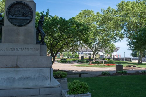 A park scene featuring a monument, trees, and a view of a stadium in the background on a clear day.