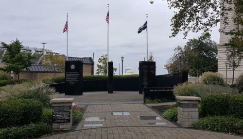 A memorial entrance with flags and black stone panels, surrounded by greenery and a paved walkway.