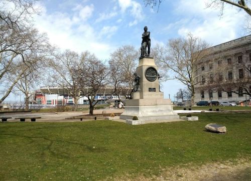 A statue on a pedestal in a park, surrounded by bare trees and grass, with a building and stadium in the background.