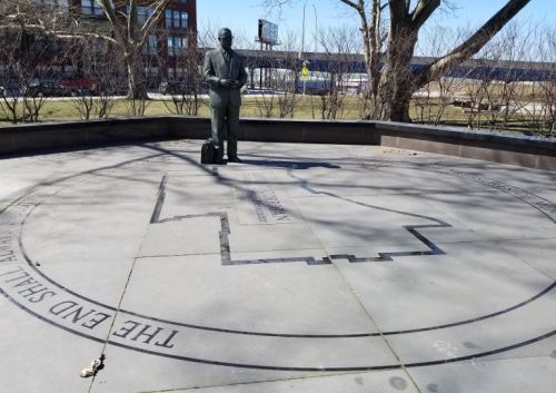 A statue of a man in a suit stands on a circular stone platform with engraved text and a map outline.