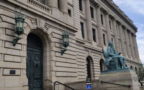 Statue of a seated figure reading, in front of a grand stone building with decorative lanterns.