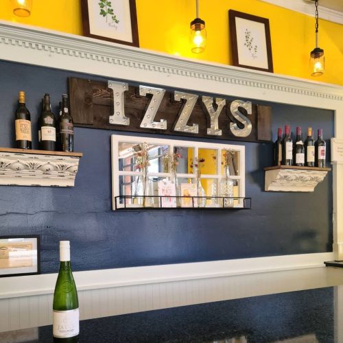A cozy wine bar with a yellow wall, shelves displaying wine bottles, and a sign that reads "IZZY'S."