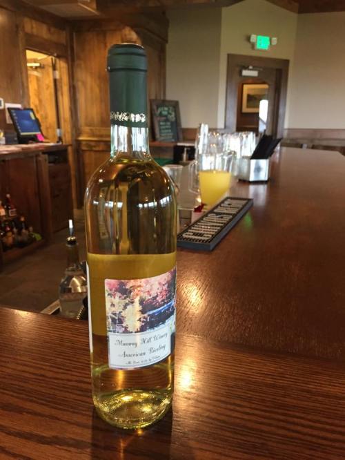 A bottle of white wine on a wooden bar, featuring a label with a scenic image and the text "Manning Hill Winery American Riesling."