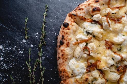 A close-up of a freshly baked pizza topped with cheese, cauliflower, and herbs on a dark slate surface.