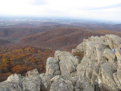 A rocky outcrop overlooks a valley filled with autumn-colored trees and distant hills under a cloudy sky.