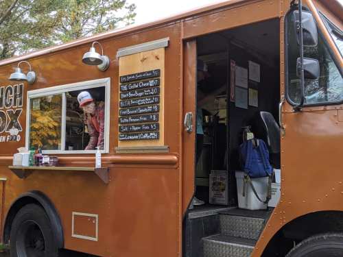 A food truck with a menu board, serving various items, and a person at the window. Trees in the background.