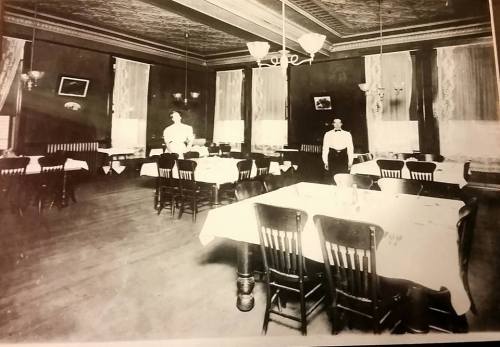 Historic dining room with tables set for guests, featuring two waitstaff in white uniforms and elegant decor.