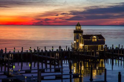 A colorful sunset over calm waters, featuring a lit-up building on a dock surrounded by boats and reflections.