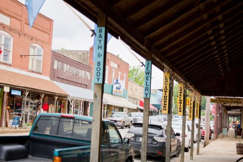 A quaint street lined with shops featuring signs for bath products, jewelry, leather goods, and more under a wooden awning.