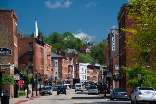 A charming street lined with historic brick buildings, shops, and greenery under a clear blue sky.