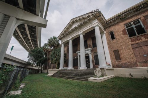 Abandoned building with tall columns, overgrown grass, and a highway overhead, under a cloudy sky.